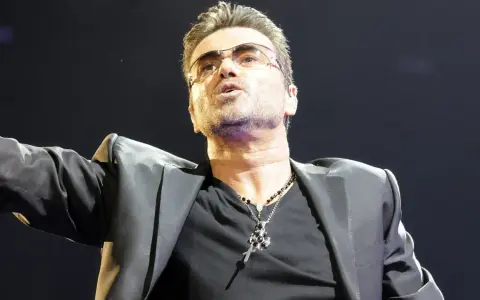 George Michael w okularach, w czarnej marynarce i z naszyjnikiem, śpiewający swoje niezapomniane utwory.