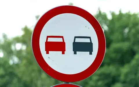 Znak drogowy: zakaz wyprzedzania. Dwa samochody, jeden czerwony, drugi czarny, stoją obok siebie.