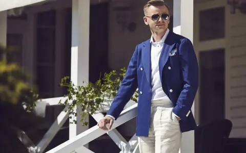 Elegancki mężczyzna w granatowej marynarce i beżowych spodniach, w stylu ivy league, stoi na werandzie.