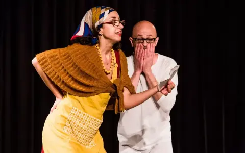 Due attori in scena, una donna con foulard e grembiule giallo, un uomo con le mani sul volto. Una scena di commedia drammatica.