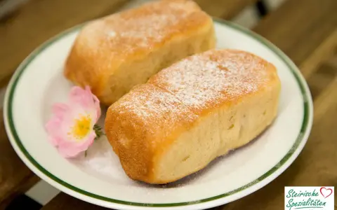 Zwei goldbraune Buchteln, bestäubt mit Puderzucker, auf einem Teller mit einer zarten rosa Blüte. Omas Rezept für Buchteln, ein Genuss!