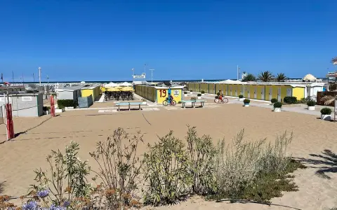 Spiaggia di Riccione con cabine gialle e blu, tavoli da ping pong e bambini in bicicletta al Bagno 19.