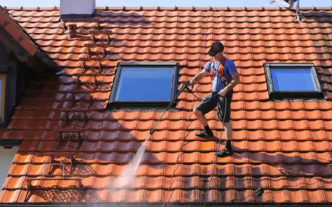 Un homme effectue le nettoyage toiture avec un nettoyeur haute pression sur un toit en tuiles rouges, sous un ciel bleu.