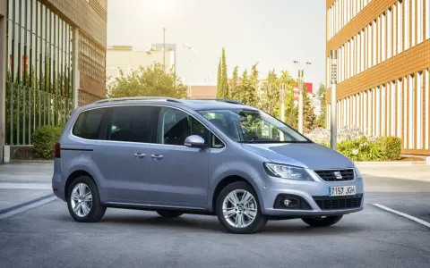 Srebrny Seat Alhambra, rodzinny van, parkuje przed nowoczesnym budynkiem z brązową elewacją.