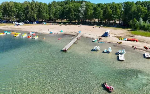 Letnia przygoda na wodzie, kolorowe rowerki wodne i kajaki czekają przy brzegu. Widok na plażę z namiotami i lasem, idealne miejsce na wypoczynek nad jeziorem Binduga 69.