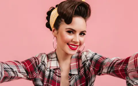 Uśmiechnięta kobieta w stylu pin-up, z burzą brązowych włosów spiętych w kok i żółtą bandaną, w kratę koszuli, robi sobie selfie.