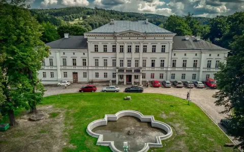 Pałac Jedlinka, biała, okazała budowla z szarym dachem, otoczona zielenią i górami. Przed pałacem fontanna i parking z samochodami.