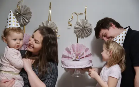 Rodzina świętuje roczek dziecka. Mama trzyma malucha, a tata podaje tort. W tle wiszą papierowe dekoracje, idealne, jak zrobić dekoracje na roczek.