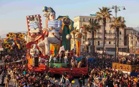 Carri allegorici e folla festante celebrano quando è Carnevale in Italia. Un clown gigante domina la scena, circondato da figure colorate e palme.