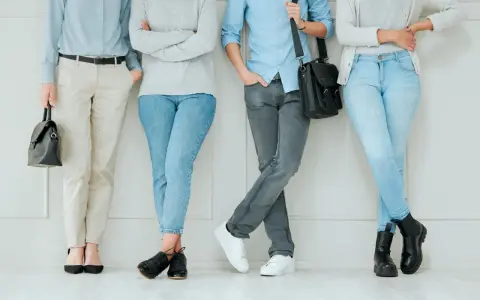 Cztery osoby w casual styl stoją przy ścianie. Jedna w jasnych spodniach i czarnych szpilkach, druga w jeansach i czarnych botkach, trzecia w szarych jeansach i białych sneakersach, czwarta w jasnych jeansach i czarnych półbutach.