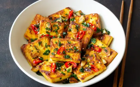 Chrupiące tofu w tempurze, posypane sezamem, chili i natką pietruszki, podane w białej misce z pałeczkami.