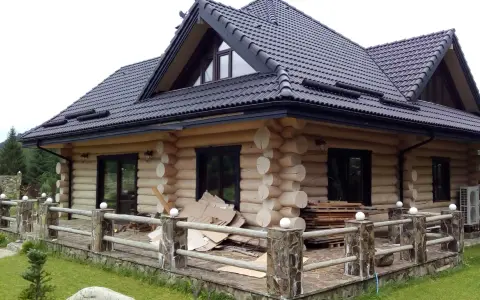 Drewniany dom z bali z czarnym dachem i tarasem, otoczony zielenią.