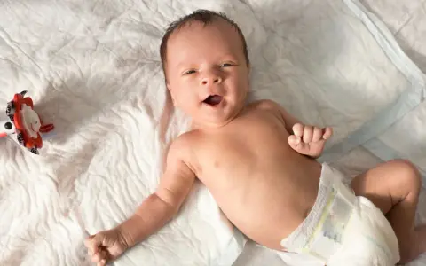 Baby mit offenem Mund, vielleicht im 6-Wochen-Schub, liegt auf einer weißen Unterlage neben einem bunten Spielzeug.