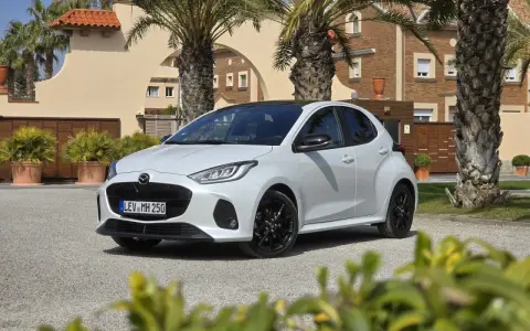 Biała Mazda 2 Hybrid zaparkowana przed budynkiem z palmami. Stylowy miejski samochód gotowy na podróż.
