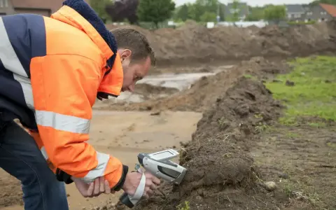 Pracownik w pomarańczowej kurtce wykonuje badanie gruntu za pomocą specjalistycznego urządzenia.