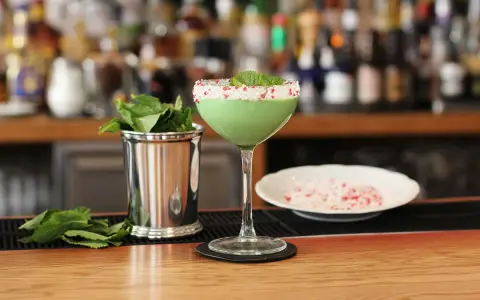 Grön drink med myntadekoration och krossad godis på kanten. Bredvid står en metallmugg med mynta. Kanske en Amarula drink?