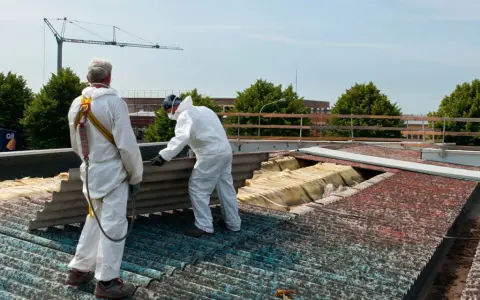 Dwóch pracowników w kombinezonach ochronnych usuwa z dachu stare płyty eternitowe. W tle widać żuraw budowlany.