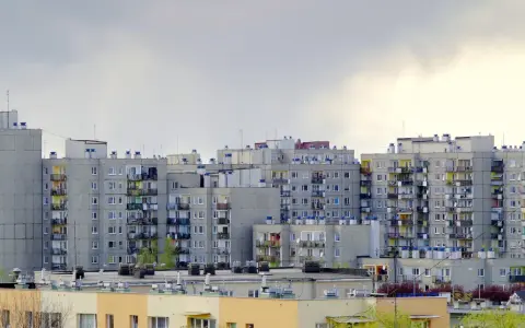 Rząd szarych bloków z wielkiej płyty, z balkonami i antenami, pod pochmurnym niebem.