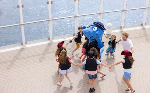 Bambini giocano in cerchio con un personaggio blu su una nave, con la costa toscana sullo sfondo.