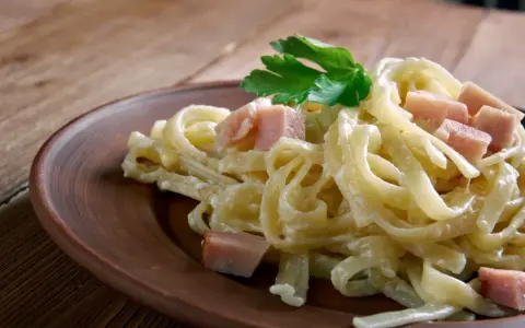 Fettuccine co to? Pyszne, kremowe fettuccine z kawałkami szynki i natką pietruszki na brązowym talerzu.