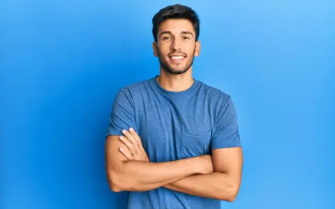 Hombre sonriente con estilos barba poco poblada, brazos cruzados, sobre fondo azul.