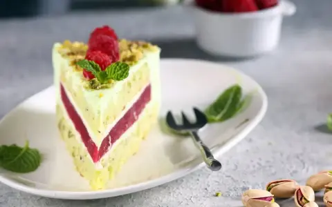 Pyszny tort pistacjowo malinowy z malinami i miętą na talerzu. Obok widoczne łuskane pistacje.