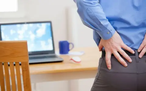 Mann mit Rückenschmerzen am Schreibtisch. Eine krankenhaustagegeldversicherung sinnvoll, um bei Krankheit abgesichert zu sein.