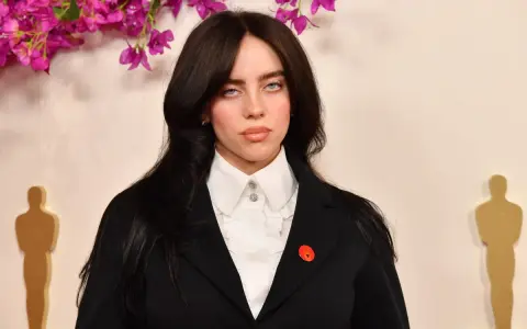 Billie Eilish z charakterystycznymi sanpaku eyes, w czarnym żakiecie i białej koszuli, na tle ozdobnych kwiatów i złotych statuetek.