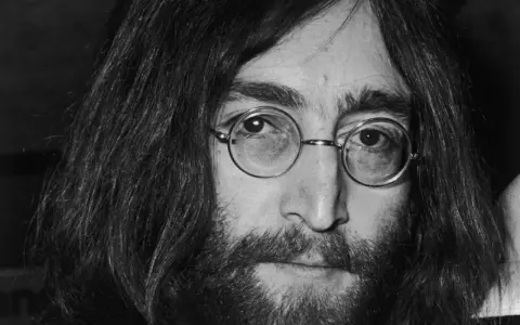 Nahaufnahme von John Lennon III. mit runden Brillengläsern und Bart.