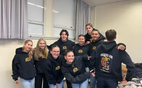 Acht lächelnde Schüler in schwarzen Hoodies mit dem Aufdruck "Volters Abi". Wie nennt man das Abitur noch?