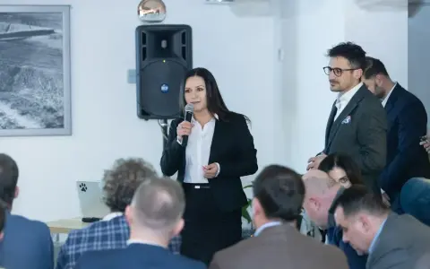 Kobieta w garniturze przemawia do grupy ludzi, obok mężczyzna w okularach.