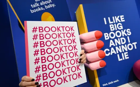 Mani che tengono un cartello con #BOOKTOK ripetuto. Sullo sfondo, un libro blu con la scritta "I LIKE BIG BOOKS AND I CANNOT LIE".