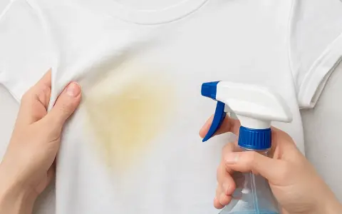 Manos aplican spray a camiseta blanca con mancha amarilla. Aprende como quitar manchas amarillas de ropa blanca fácilmente.