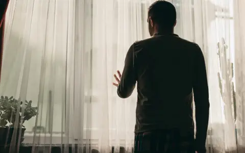 Mężczyzna stoi przy oknie, zasłoniętym przezroczystą tkaniną. Jego dłoń opiera się o materiał, jakby szukał kontaktu ze światem zewnętrznym. Obraz symbolizuje schizofrenia objawy, izolację i trudności w komunikacji.