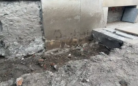 Widoczne ślady osiadania budynku i pęknięcia na cokole. Uszkodzony fragment fundamentu, ziemia i gruz wokół.