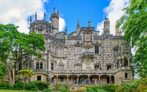 The Quinta da Regaleira estate in Sintra, Portugal, features a stunning palace with intricate stonework and ornate balconies, surrounded by lush greenery under a blue sky.