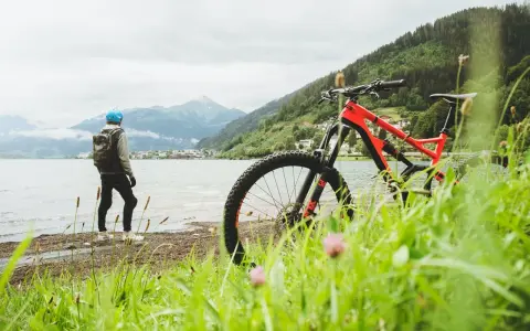 En cyklist med hjälm och ryggsäck står vid en sjö med berg i bakgrunden. En mountainbike står redo för äventyr på mtb leder.