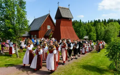 En procession av musiker i folkdräkter, som spelar fiol och gitarr, går förbi en röd träkyrka. Många människor i traditionella kläder, vilka firar midsommar, följer efter.