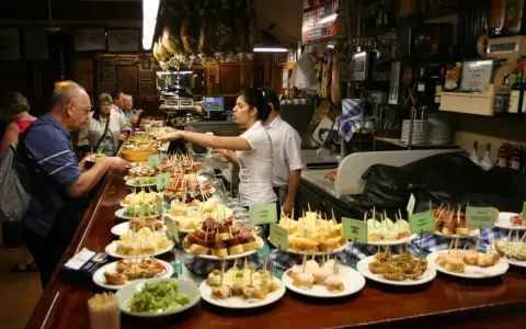 Tapas bar pełen ludzi wybierających przekąski. Na ladzie mnóstwo małych kanapeczek i innych przysmaków.