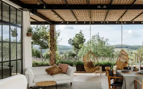 Terraza minimalista con sofá blanco, sillas colgantes de mimbre y vistas a un paisaje verde.