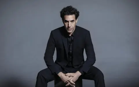Sacha Baron Cohen, känd för sina kontroversiella filmer, sitter i en mörk kostym med händerna knäppta.