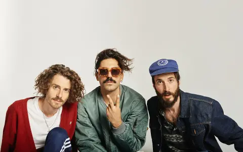 Tres hombres posan, uno con cárdigan rojo, otro con gafas de sol y chaqueta bomber, y el tercero con gorra azul. Parecen un grupo de música, quizás el cantante sexy de Zebras.