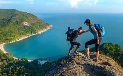 Due escursionisti si aiutano a salire su una scogliera con vista su una baia tropicale. Questo è il trekking cos'è: avventura e collaborazione.
