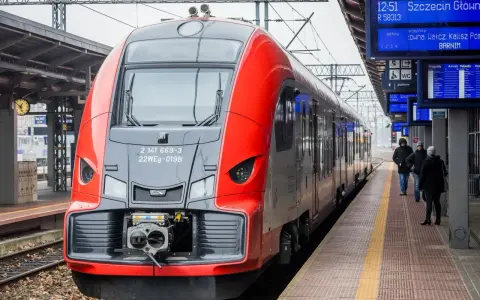 Czerwono-szary pociąg stoi na peronie, obok tablic informacyjnych z rozkładem jazdy.