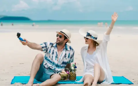 Para na plaży nagrywa filmik kamerką GoPro, a jego partnerka macha do obiektywu. Idealne wakacje pod gruszą!