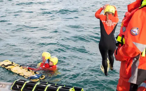 Sauvetage en mer : une sauveteuse en combinaison noire et orange se prépare à plonger. Deux personnes en gilets de sauvetage et casques jaunes flottent près d'une civière. Communication VHF et MMSI essentielle.