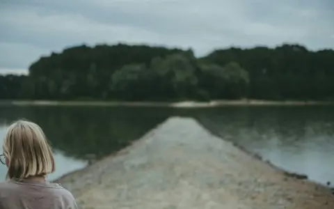 Dziewczyna z blond włosami patrzy na spokojną taflę jeziora, czując uczucie pustki.