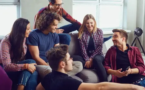 Grupa przyjaciół śmieje się, siedząc na kanapie. Wyglądają, jakby grali w grę, może nawet w Familiadę, odpowiadając na zabawne pytania.