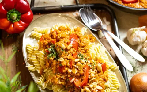 Fusilli mit einer herzhaften bolognese mit roten linsen, garniert mit Erdnüssen und Petersilie. Daneben liegen eine rote Paprika und Knoblauch.