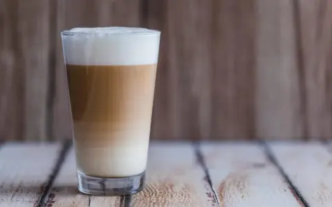 Ein hoher Latte Macchiato mit Mandelmilch, geschichtet in drei Farben, steht auf einem Holztisch.